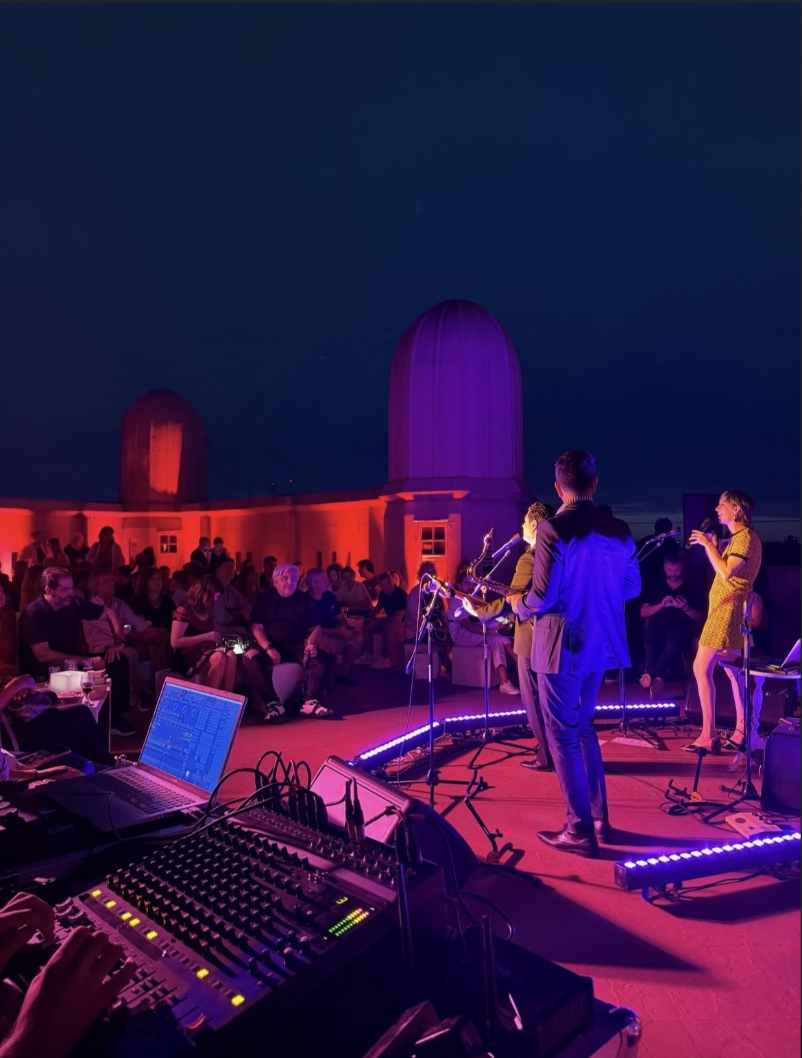 Banda tocando en una terraza con una cúpula histórica de fondo, iluminación roja y violeta